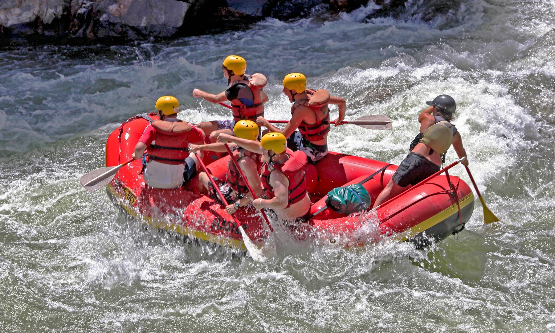 A group of people in life jackets and helmets paddling down a section of white water rapids in a red raft.