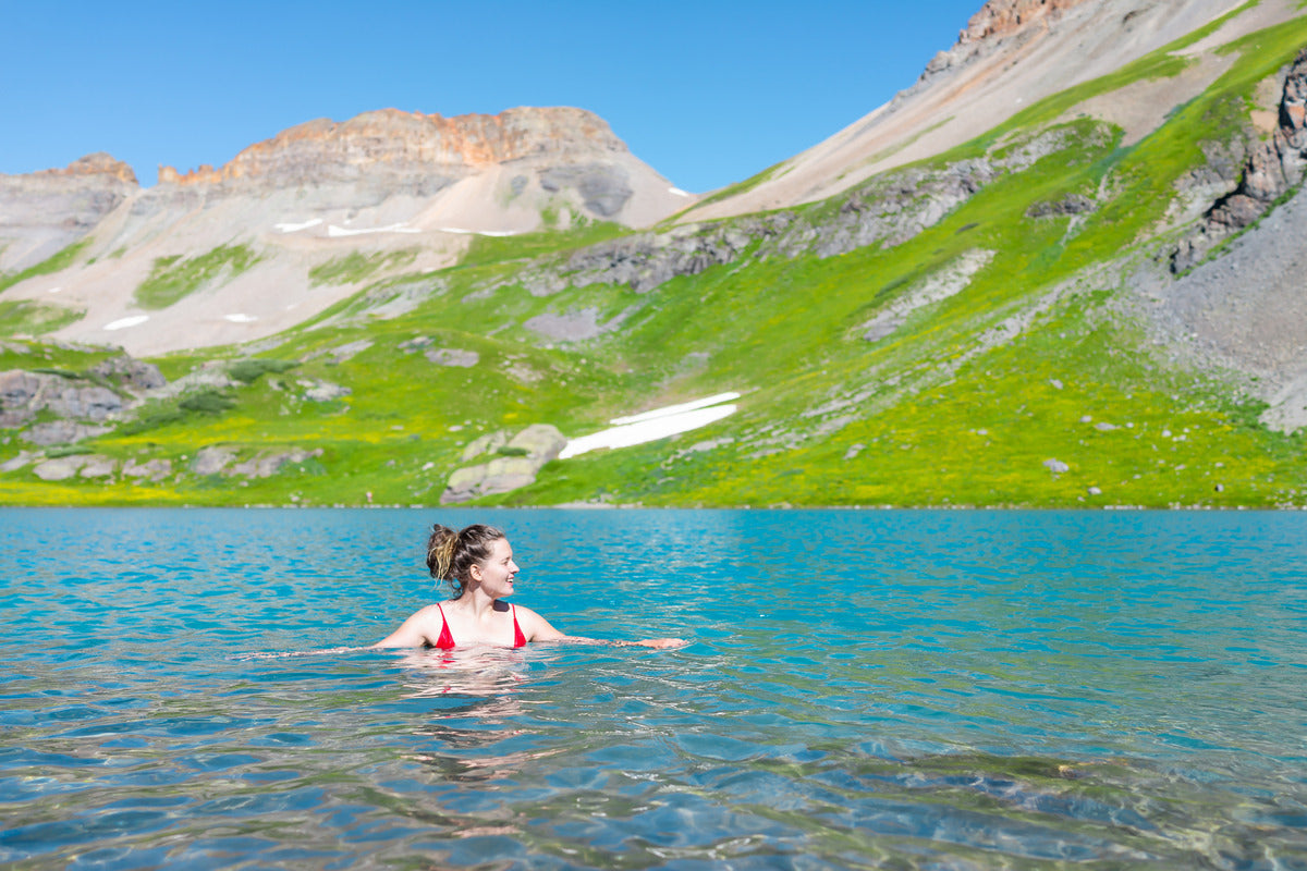 A woman in a red swimsuit treading water in the middle of a vibrant mountain lake surrounded by lush green slopes.