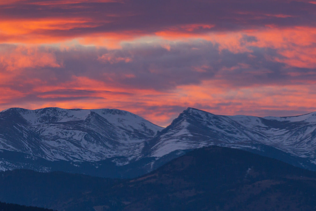 A sunset over the snow-capped Rocky Mountains. The sky is full of clouds reflecting the red and gold of the setting sun.