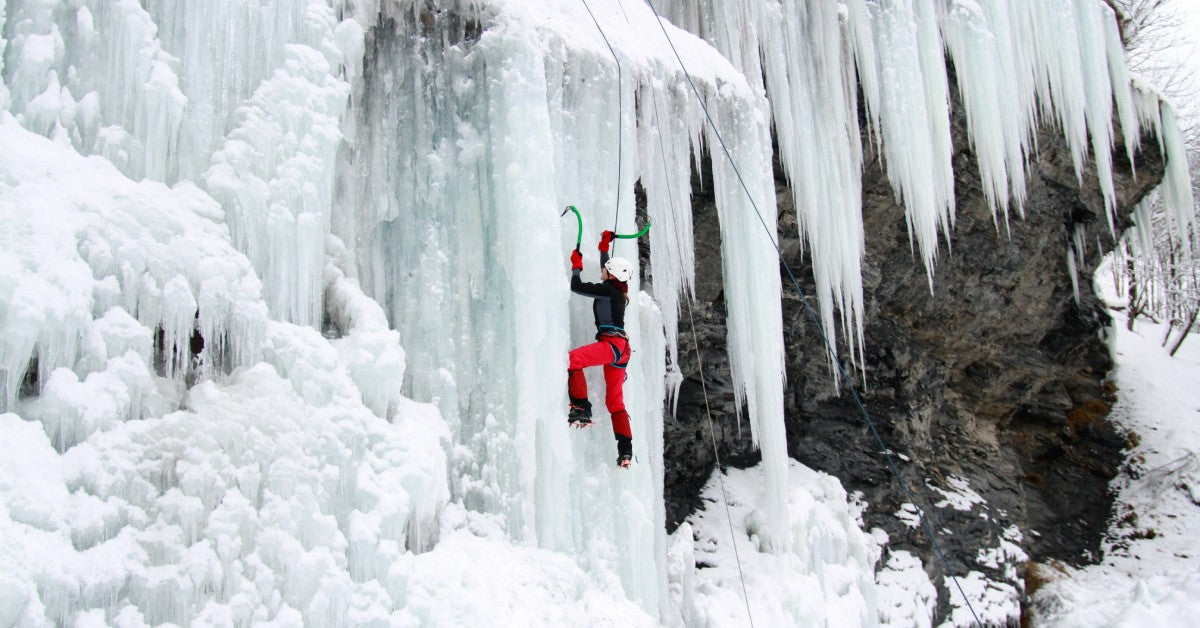 A woman wearing a helmet, gloves, and black and red sports gear using a harness and picks to climb a frozen waterfall.