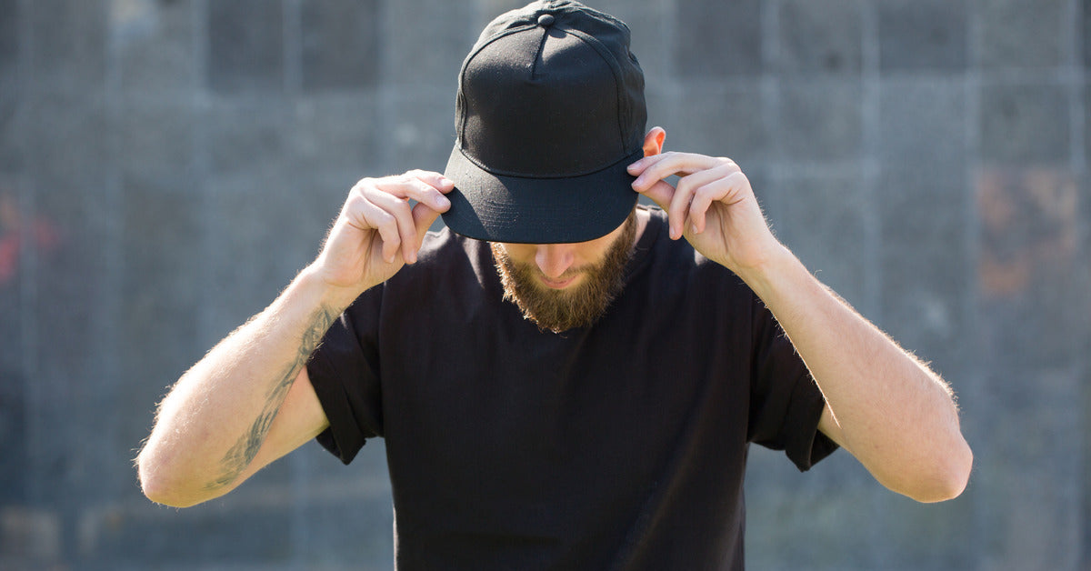 A man in a black t-shirt and a beard wearing a black baseball cap. He is reaching up and adjusting the hat's brim.