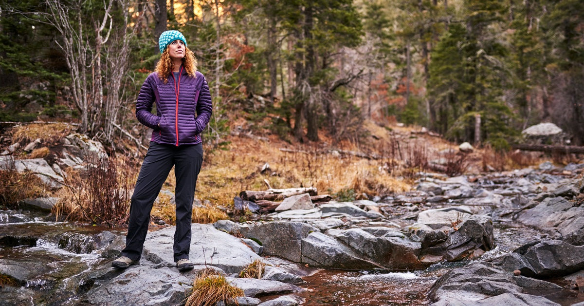 A woman wearing a beanie and a jacket stands at the edge of a creek in a forest. She is looking off into the distance.